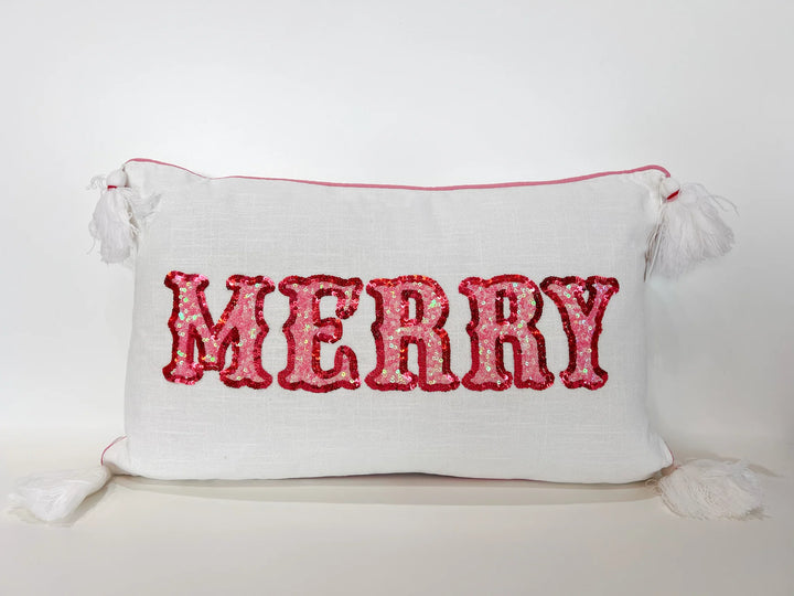 White fabric lumbar pillow featuring the word MERRY in shimmering pink and red sequins, accented with pink-piped edges and four large white corner tassels against a neutral white background