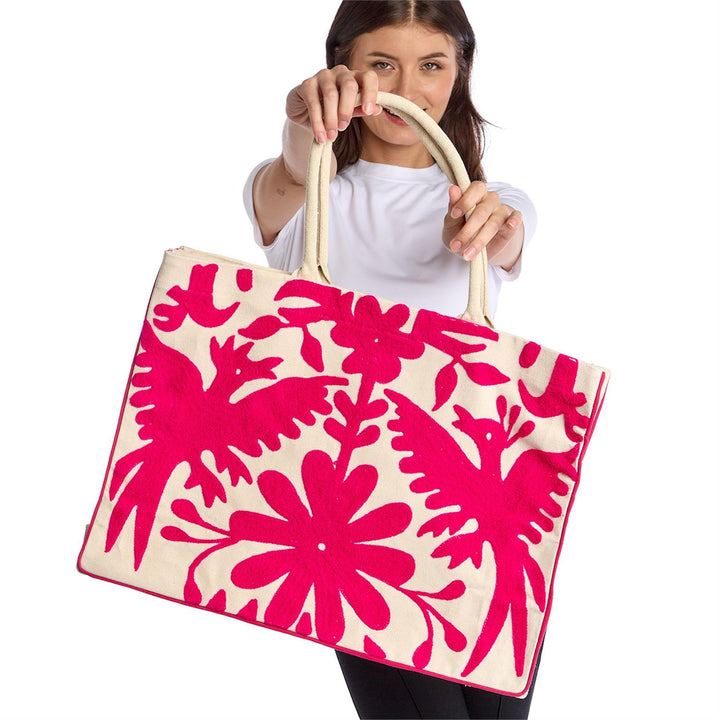Otomi Tenango hand-embroidered cotton tote bag held by a woman against a white studio background, featuring vibrant fuchsia stylized birds and large floral patterns with thick cream-colored rope handles
