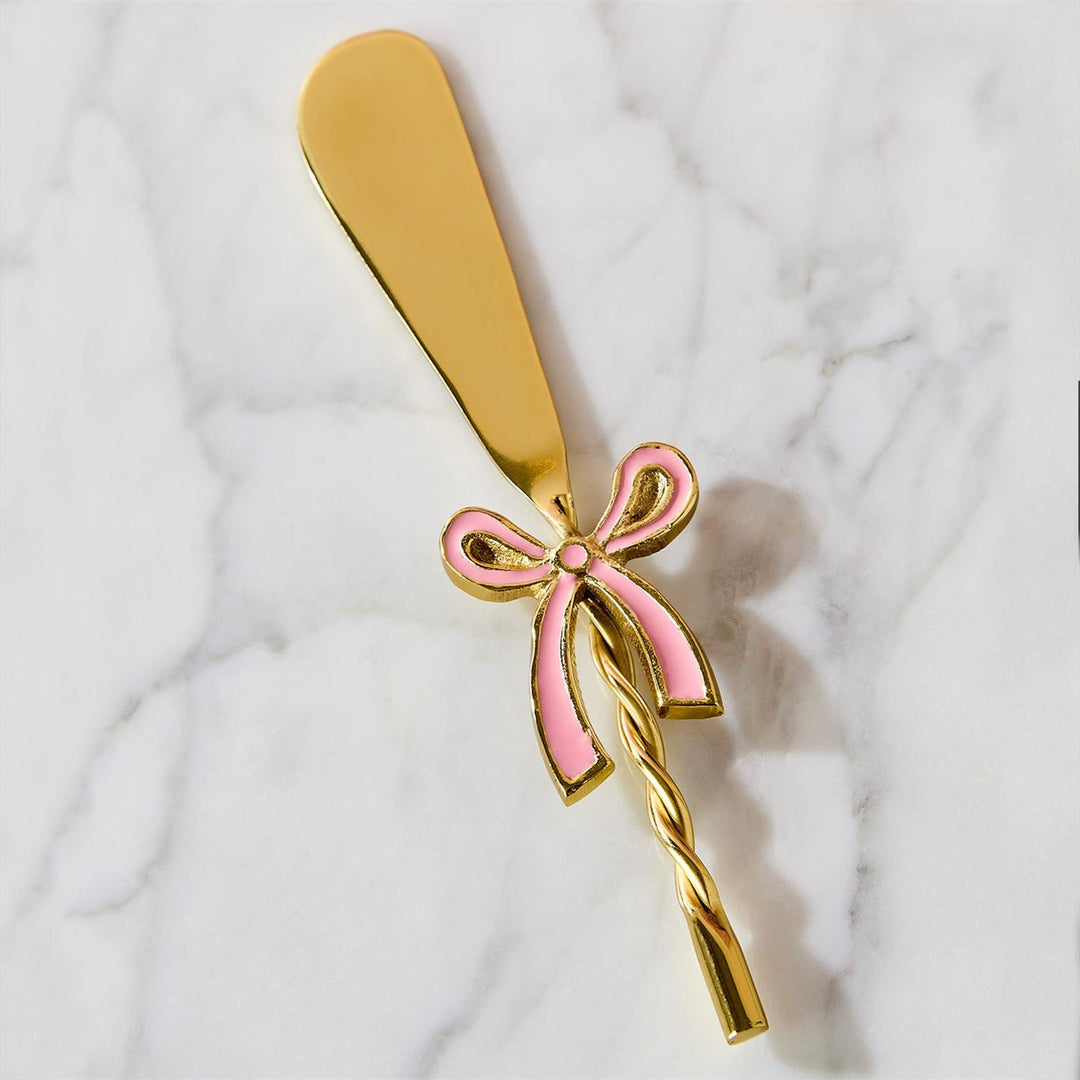 Gold-toned metal butter spreader featuring a decorative pink enamel bow and a twisted handle resting on a white veined marble surface with a wide flat blade and reflective polished finish