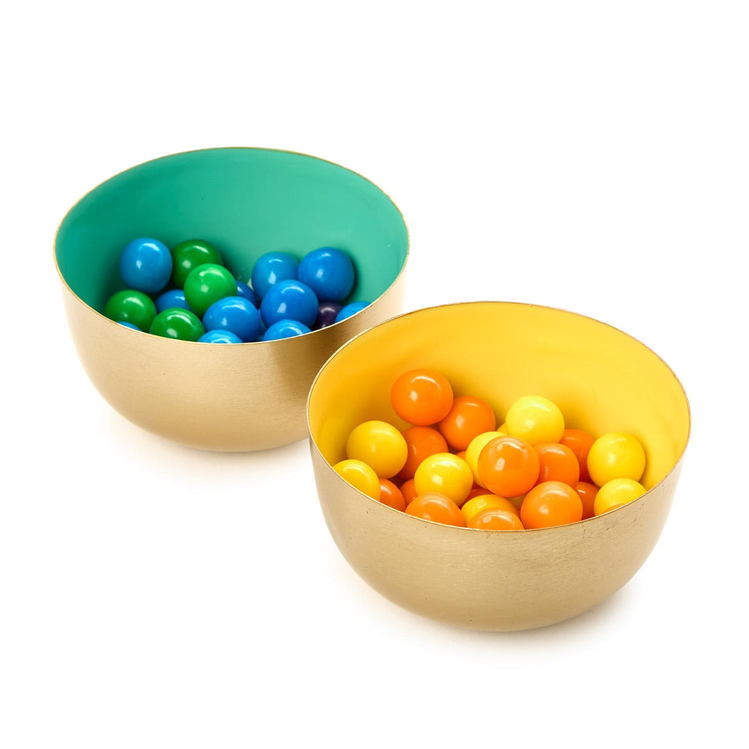 Metal and enamel decorative bowls featuring brushed gold exteriors and vibrant turquoise and yellow interior linings filled with glossy blue, green, and orange gumballs on a white studio background