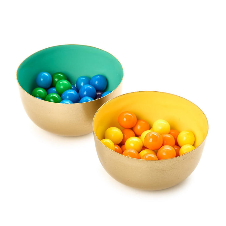 Metal and enamel decorative bowls featuring brushed gold exteriors and vibrant turquoise and yellow interior linings filled with glossy blue, green, and orange gumballs on a white studio background