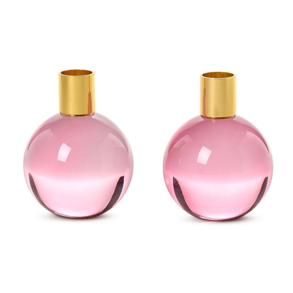 Hand-blown crystal candle holders by Anna Ehrner for Kosta Boda featuring two translucent pink spherical bases with polished gold-toned brass collars and subtle reflective highlights on a plain white background