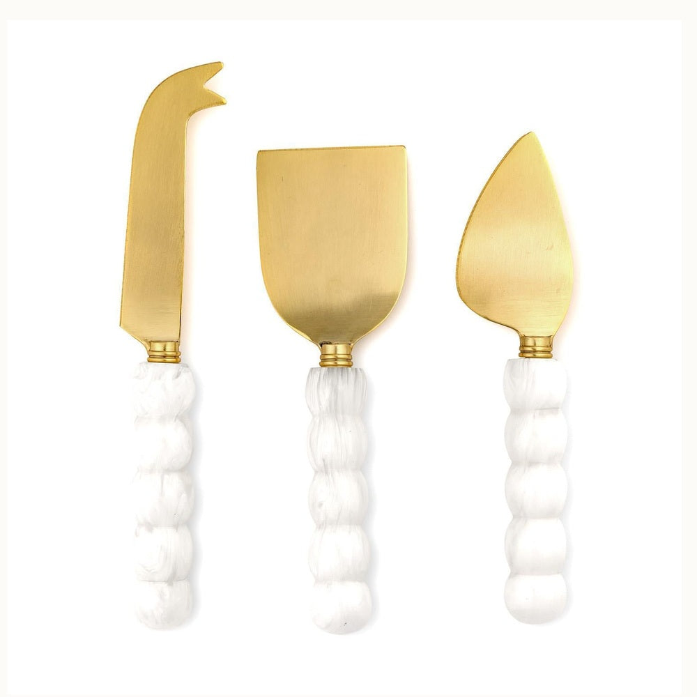 Gold-finished stainless steel three-piece cheese knife set featuring a flat spade, fork-tipped spear, and pointed almond knife with white marbled bobbin-style handles and a unique scalloped texture against a plain white background
