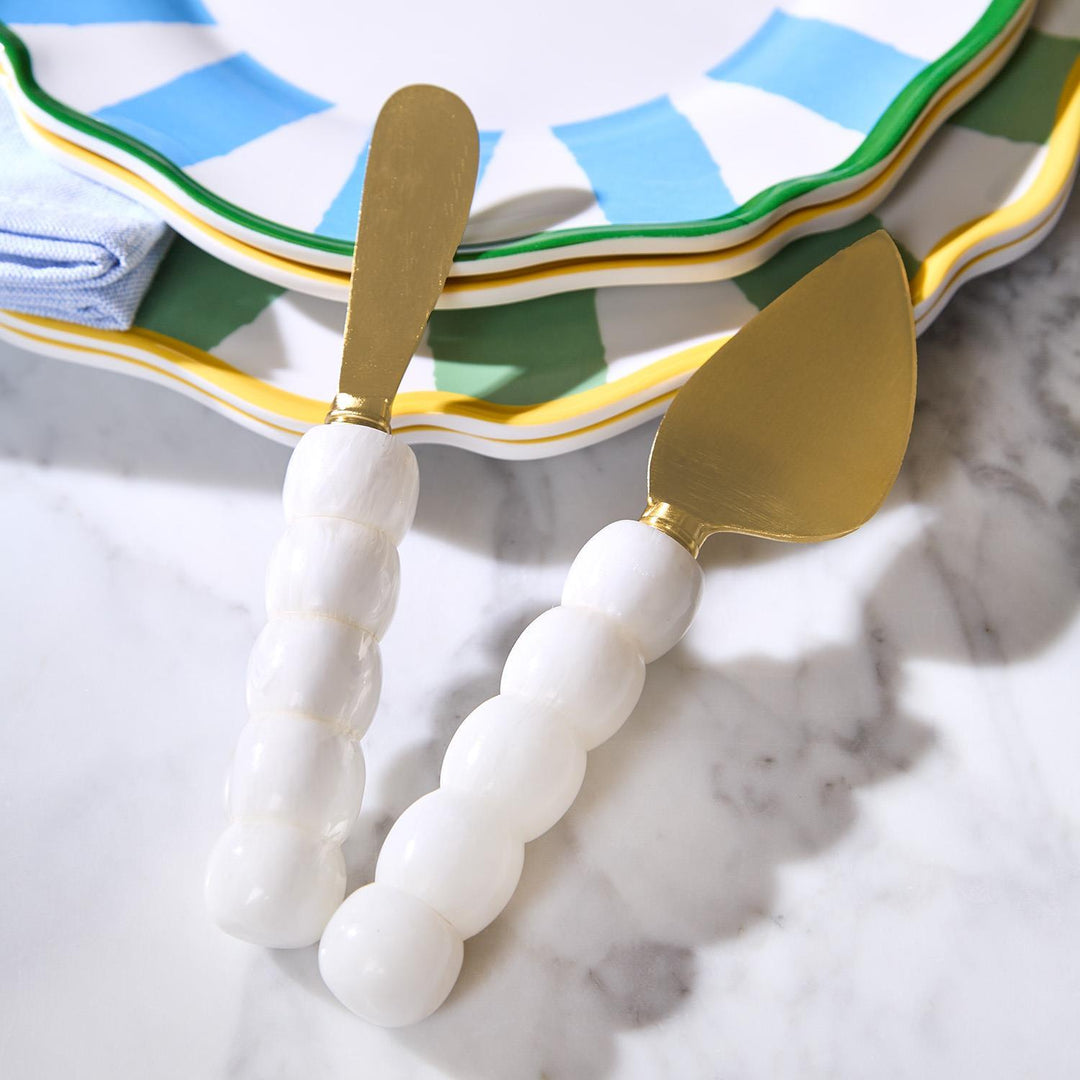 Lifestyle product photography of gold cheese spreaders with white bulbous handles resting on a marble surface beside stacked scalloped plates with vibrant blue and green stripes