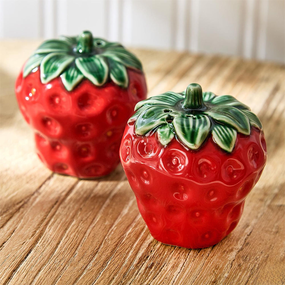 Glossy ceramic strawberry salt and pepper shakers sit on a rustic wooden table featuring a vibrant red glaze, embossed seed textures, and sculpted green calyx stems with small shaker holes