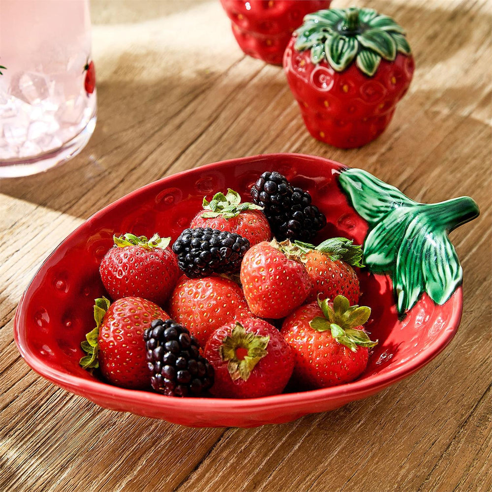 Photograph of fresh red strawberries and dark blackberries in a glossy strawberry-shaped ceramic bowl with textured green leaf handles on a rustic wooden table next to a cold pink beverage under natural light
