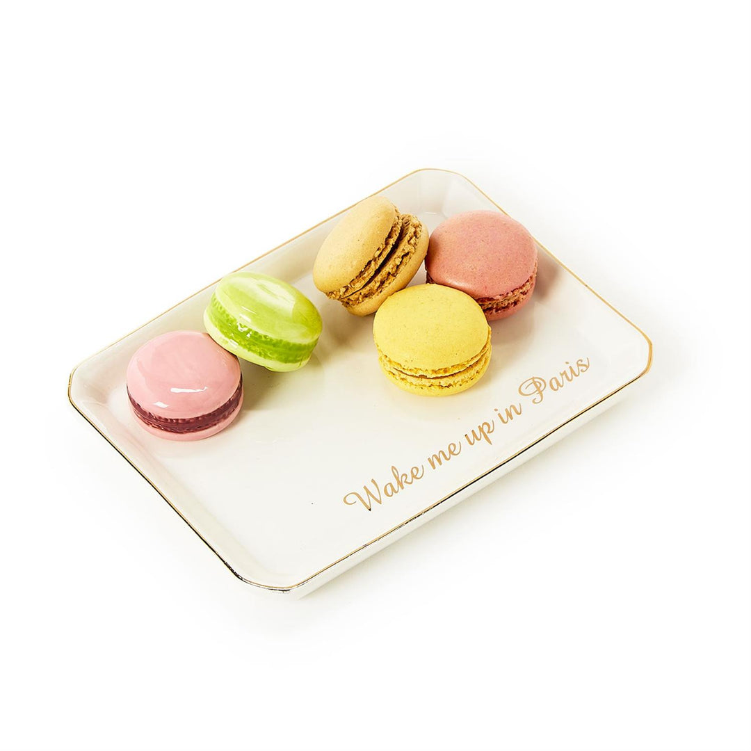 Still life photograph of five colorful macarons in pastel pink, lime green, and yellow arranged on a gold-rimmed white ceramic tray inscribed with the elegant gold script Wake me up in Paris