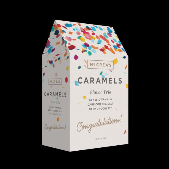Product photograph by McCrea's of a white gable-top caramel flavor trio gift box against a solid black background featuring multi-colored confetti and elegant cursive text reading Congratulations