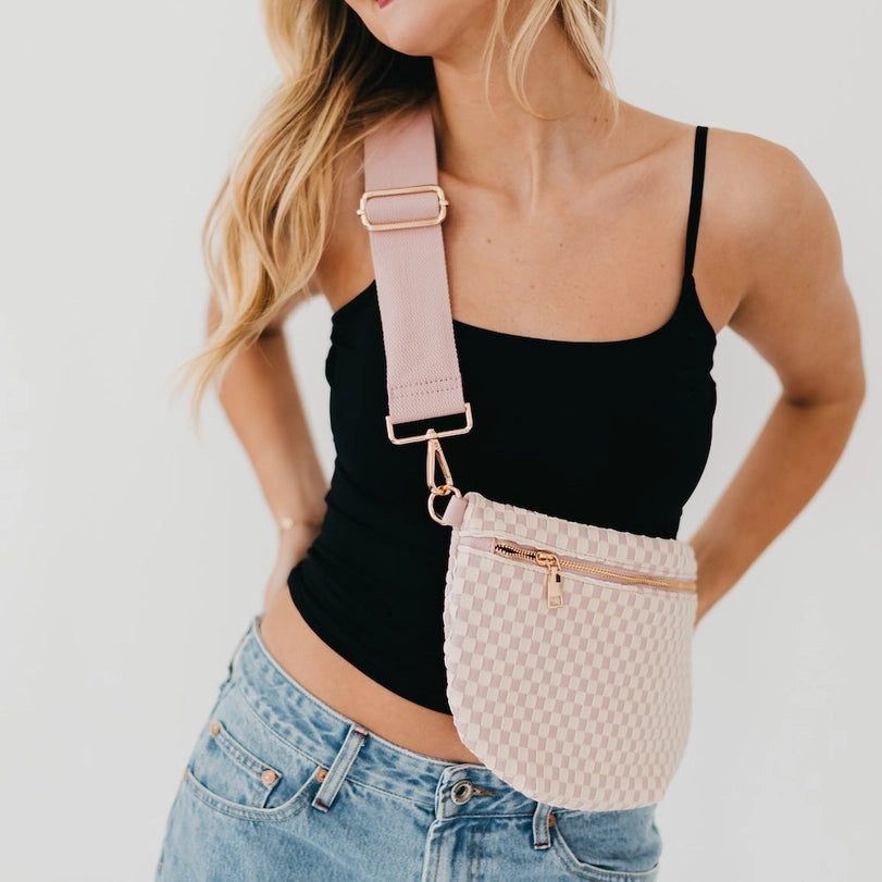 Lifestyle photograph of a woman wearing a pink checkered crossbody fanny pack against a white studio background, highlighting its wide adjustable strap, gold-toned metal hardware, and light-wash denim jeans