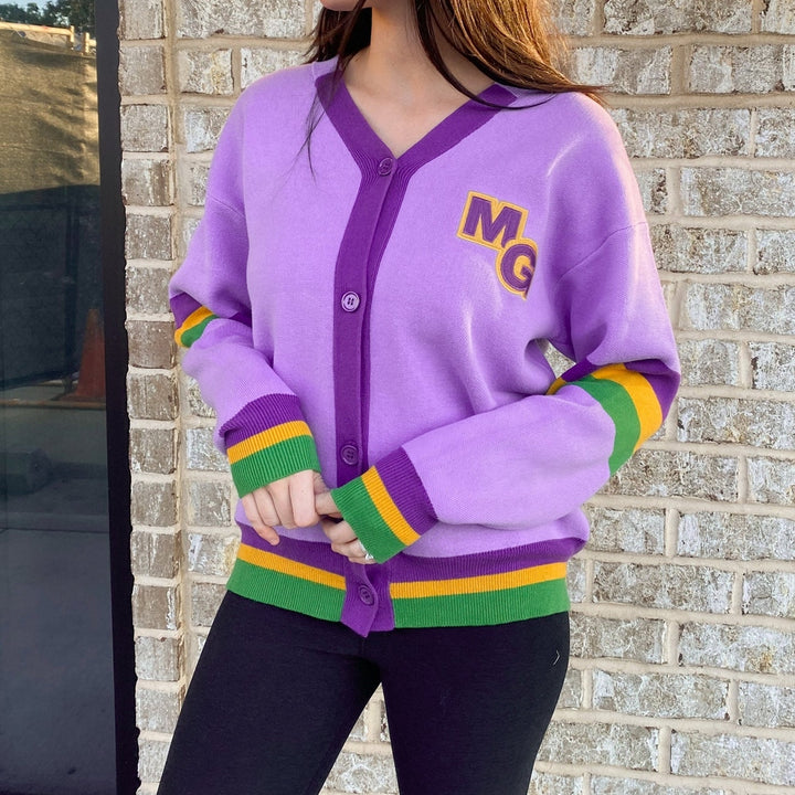 Photograph of a person wearing a lavender varsity cardigan featuring a yellow MG patch and green and gold striped cuffs while standing in front of a light brick wall