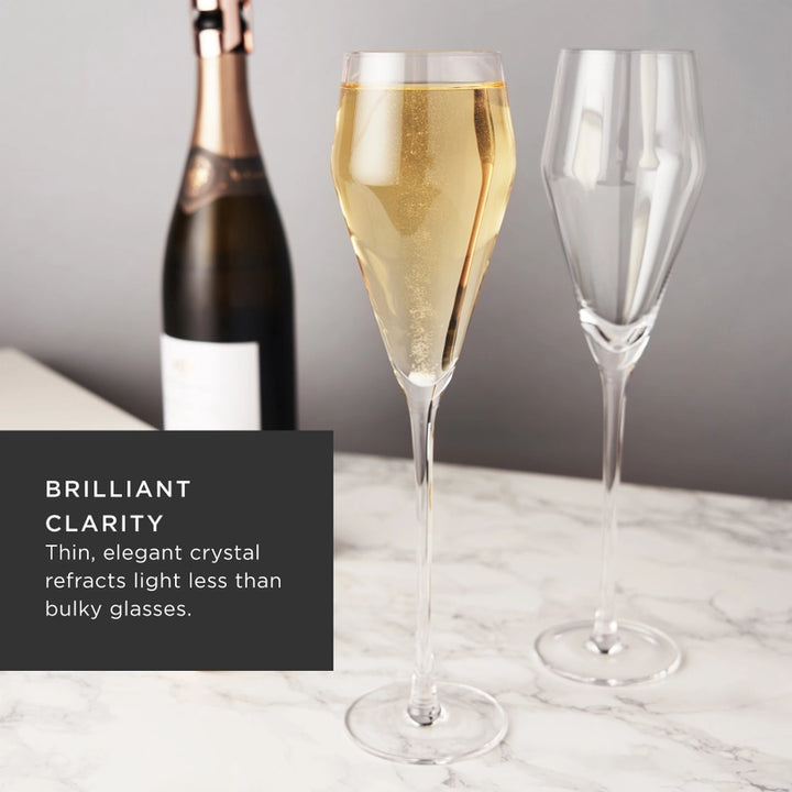 Commercial product photograph of two elegant crystal champagne flutes on a white marble surface featuring one glass filled with bubbly golden liquid and a champagne bottle with rose gold foil in the background