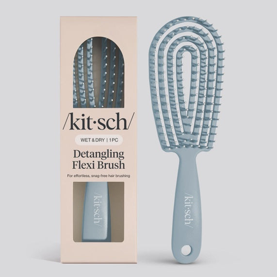 Commercial product photography by Kitsch displays a dusty blue Detangling Flexi Brush and its beige packaging on a grey studio background, featuring a curved flexible vented head, soft plastic bristles, and the prominent Kitsch logo