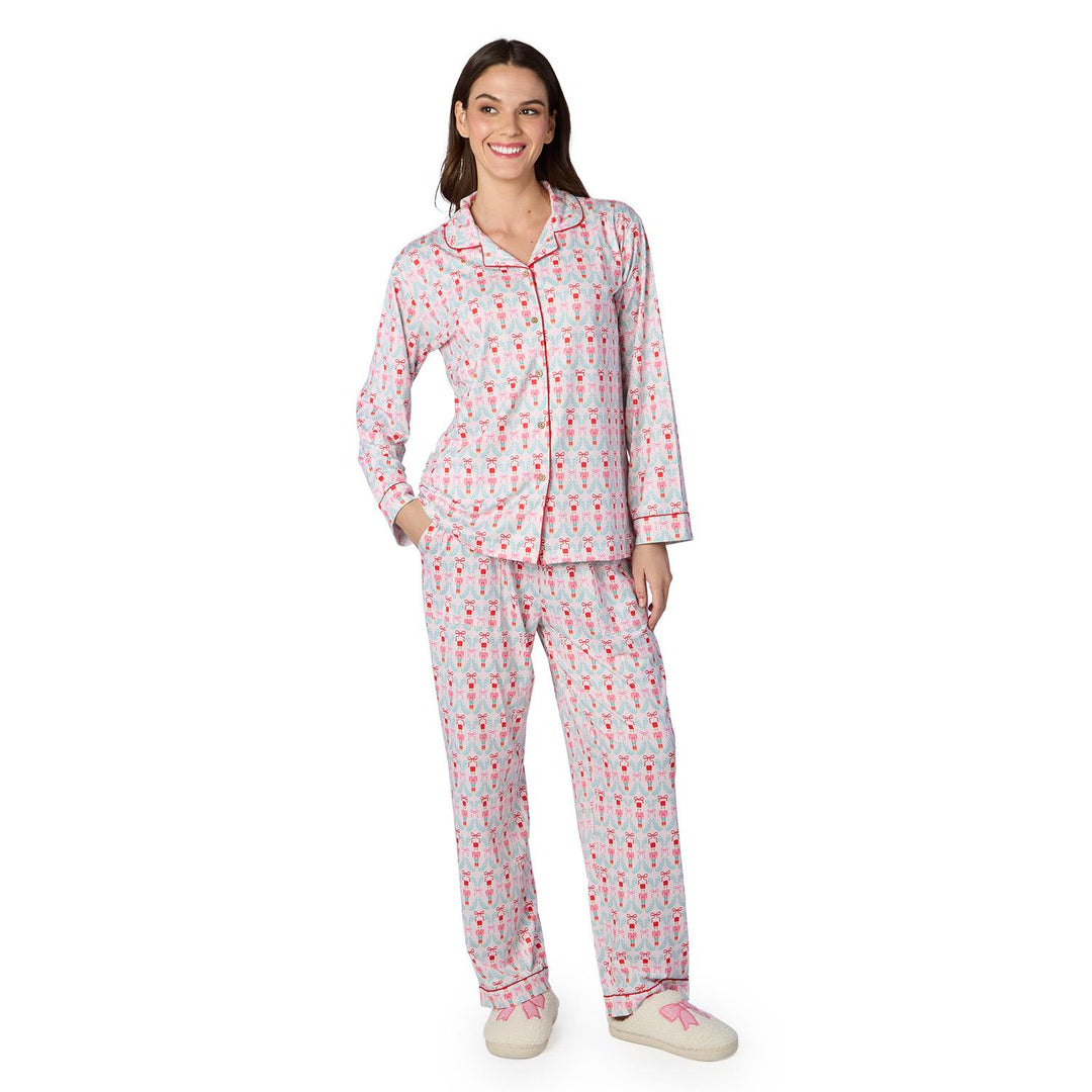 Digital photograph of a smiling woman wearing a light blue pajama set with a nutcracker and pink bow pattern featuring red piped trim and matching slippers against a white studio background