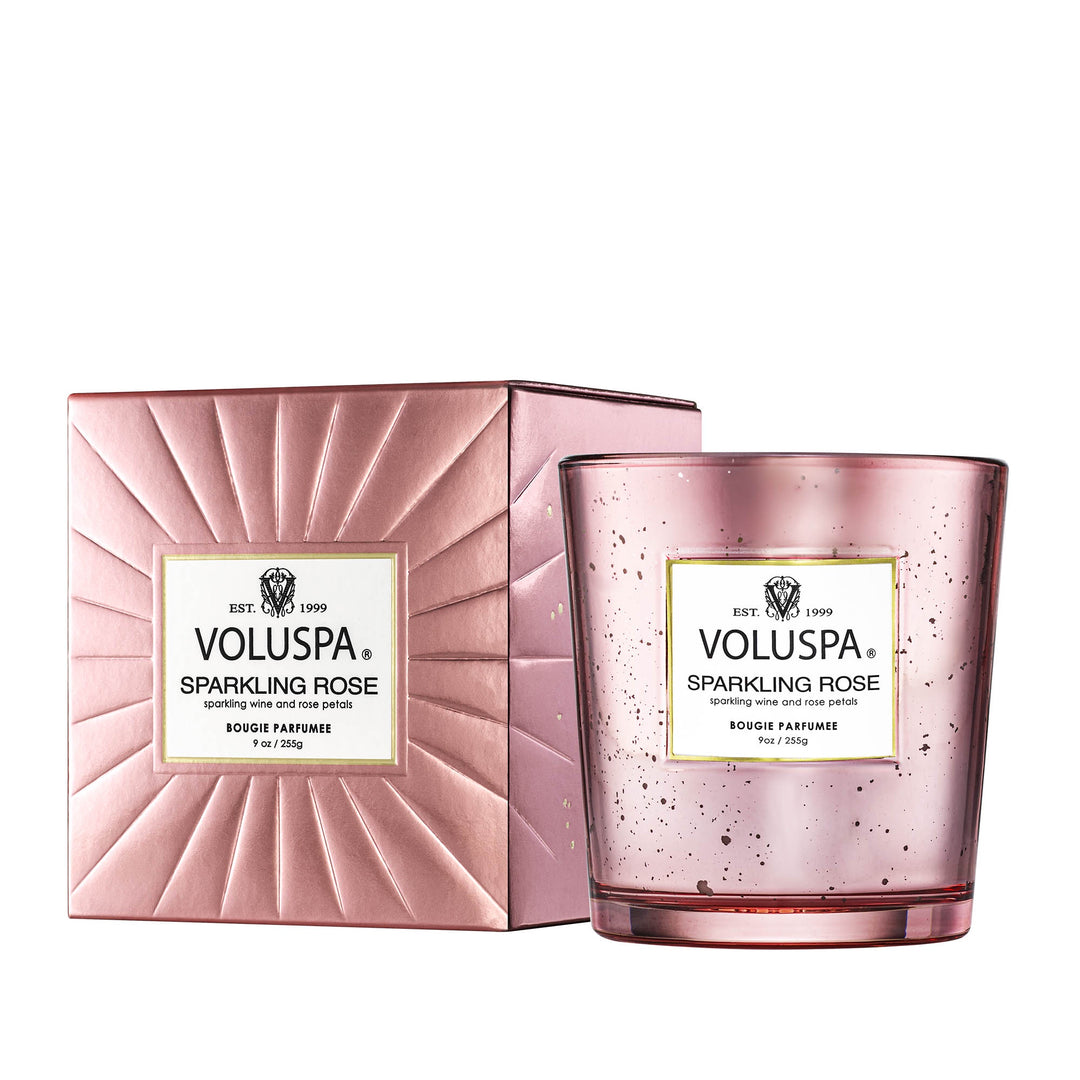 Product photograph by Voluspa of a Sparkling Rose scented candle and gift box featuring a metallic pink sunburst texture, a prominent logo label, and a speckled mercury glass jar