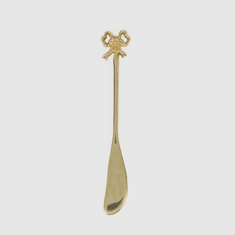 Gold-plated metal decorative butter spreader centered on a white background featuring an intricate ribbon bow handle, a long slender neck, and a polished asymmetrical blade with a reflective finish