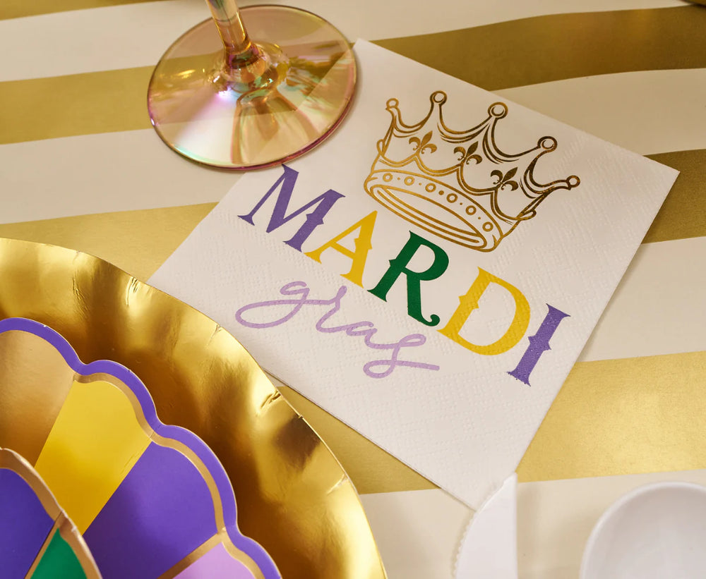 Photograph of a festive Mardi Gras party table setting featuring a white napkin with a metallic gold crown and colorful script beside metallic scalloped plates on a gold-and-white striped tablecloth