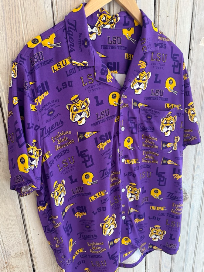 Short-sleeved button-down shirt featuring an allover LSU Fighting Tigers graphic print with Mike the Tiger mascot heads, yellow football helmets, and collegiate pennants on a vibrant purple background with a single chest pocket