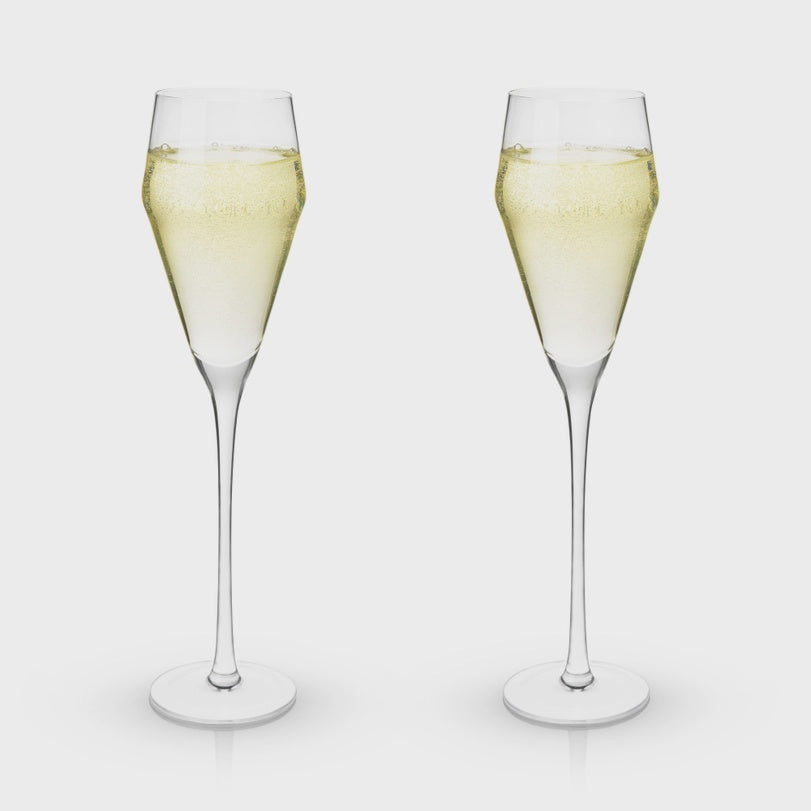 Product photograph of two modern tulip-style crystal champagne flutes filled with sparkling pale gold wine featuring tall slender stems and fine rising bubbles against a neutral grey background