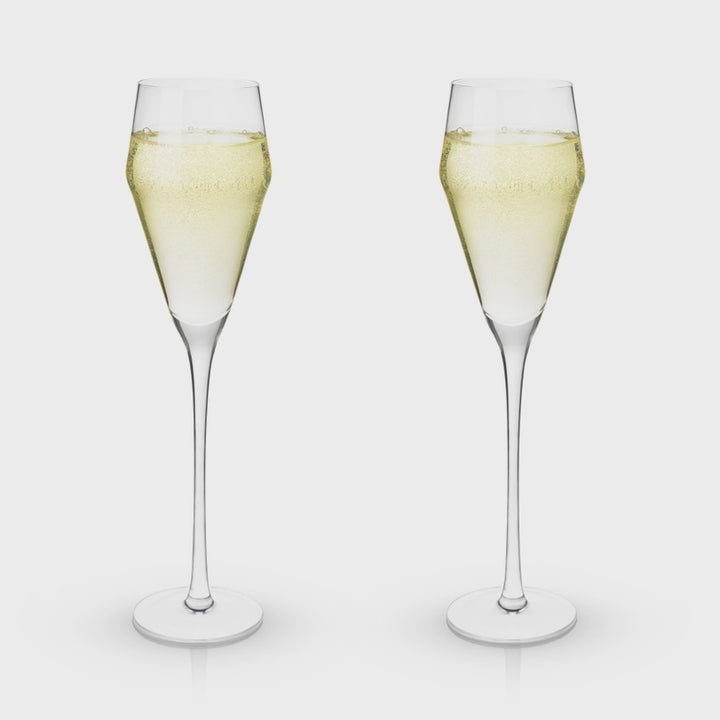 Product photograph of two modern tulip-style crystal champagne flutes filled with sparkling pale gold wine featuring tall slender stems and fine rising bubbles against a neutral grey background
