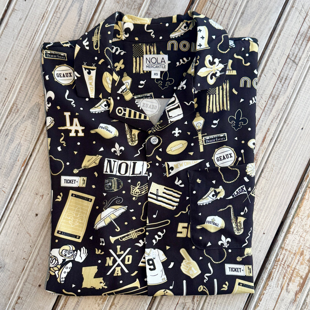 Button-down shirt by NOLA Mercantile showcases a New Orleans themed pattern on black fabric featuring iconic Bourbon Street signs, a Louisiana state map, and vibrant gold fleur-de-lis symbols alongside saxophone and football motifs