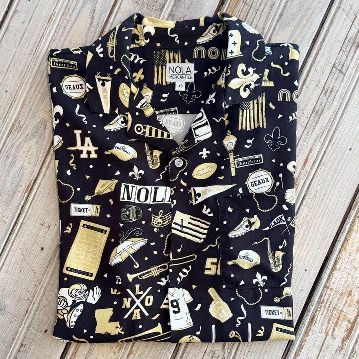 Button-down shirt by NOLA Mercantile showcases a New Orleans themed pattern on black fabric featuring iconic Bourbon Street signs, a Louisiana state map, and vibrant gold fleur-de-lis symbols alongside saxophone and football motifs