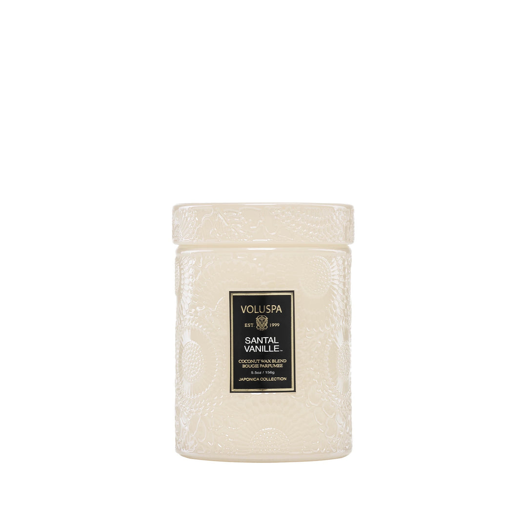 Glass jar candle by Voluspa featuring Santal Vanille from the Japonica collection centered on a white background with intricate cream-colored embossed floral patterns, a matching glass lid, and a black and gold label