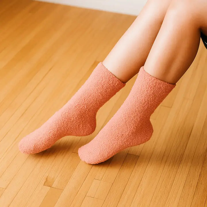 Photograph of a person’s legs wearing peach-colored plush fuzzy socks resting on a light-toned polished hardwood floor