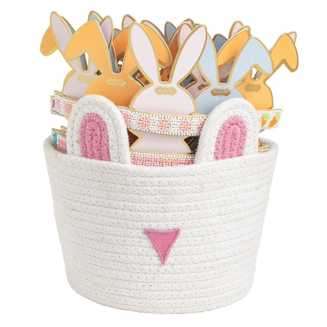 Product photograph by Mud Pie of a white woven rope bunny basket with a pink embroidered nose filled with decorative headbands featuring pastel rabbit silhouettes and intricate floral beadwork designs