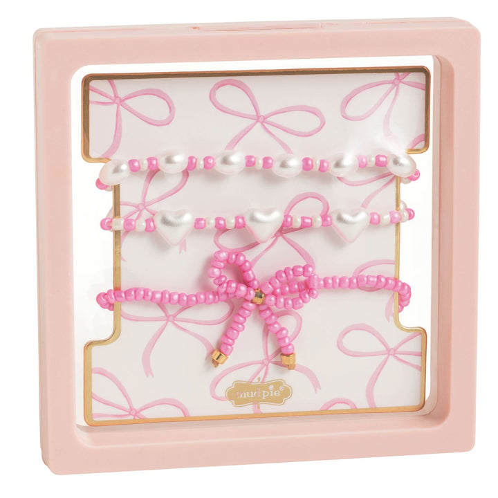 Beaded jewelry set by Mud Pie featuring three pink and white bracelets displayed in a pink frame with iridescent faux pearls, heart-shaped charms, and a large pink beaded bow with gold tips