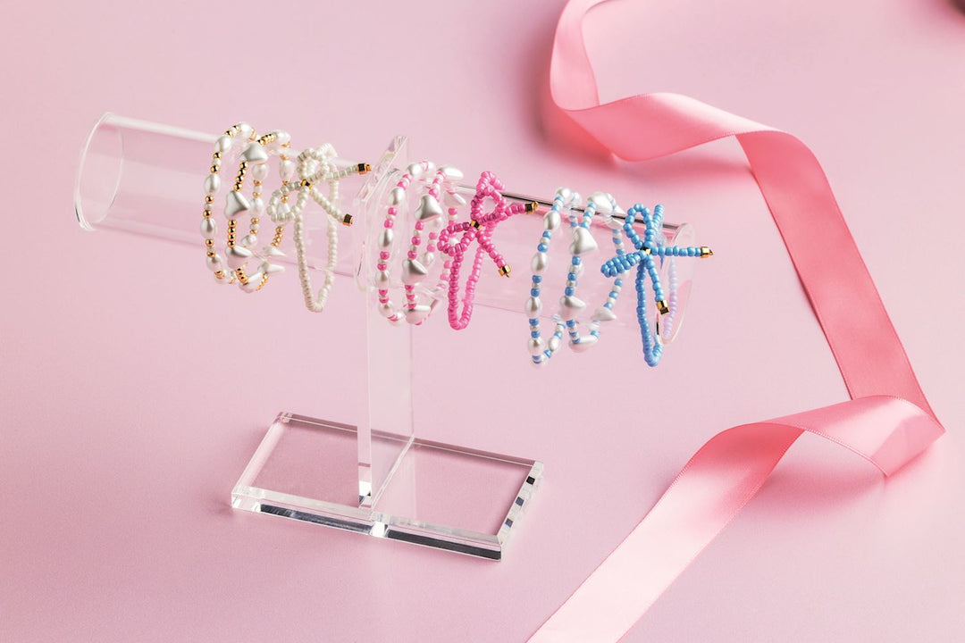 Commercial product photograph of handmade beaded bracelets featuring white heart charms and colorful beaded bows displayed on a transparent acrylic T-bar stand alongside a shimmering pink satin ribbon on a soft pastel background
