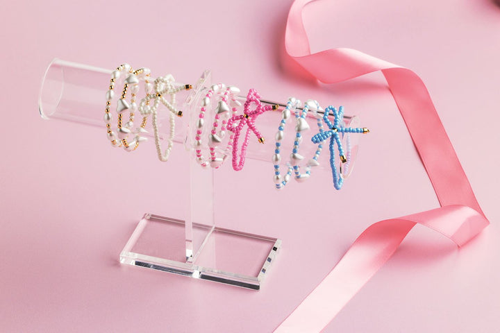 Commercial product photograph of handmade beaded bracelets featuring white heart charms and colorful beaded bows displayed on a transparent acrylic T-bar stand alongside a shimmering pink satin ribbon on a soft pastel background