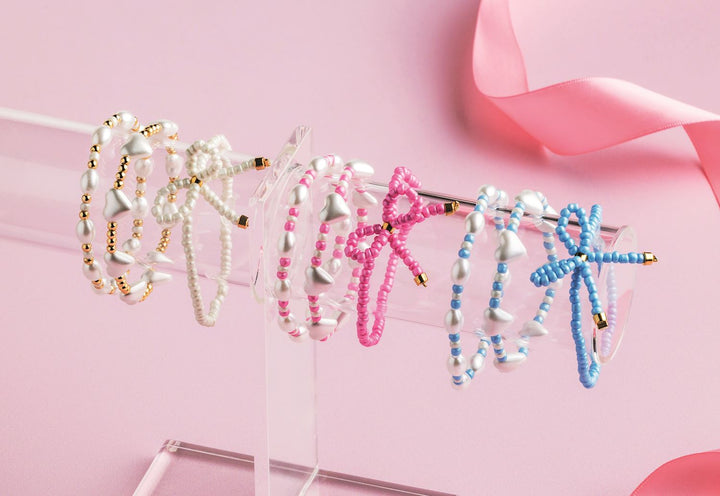 Photograph of handmade beaded bracelets on a clear acrylic display stand featuring white heart-shaped pearls, vibrant pink and blue beaded bows, and a soft pink ribbon background