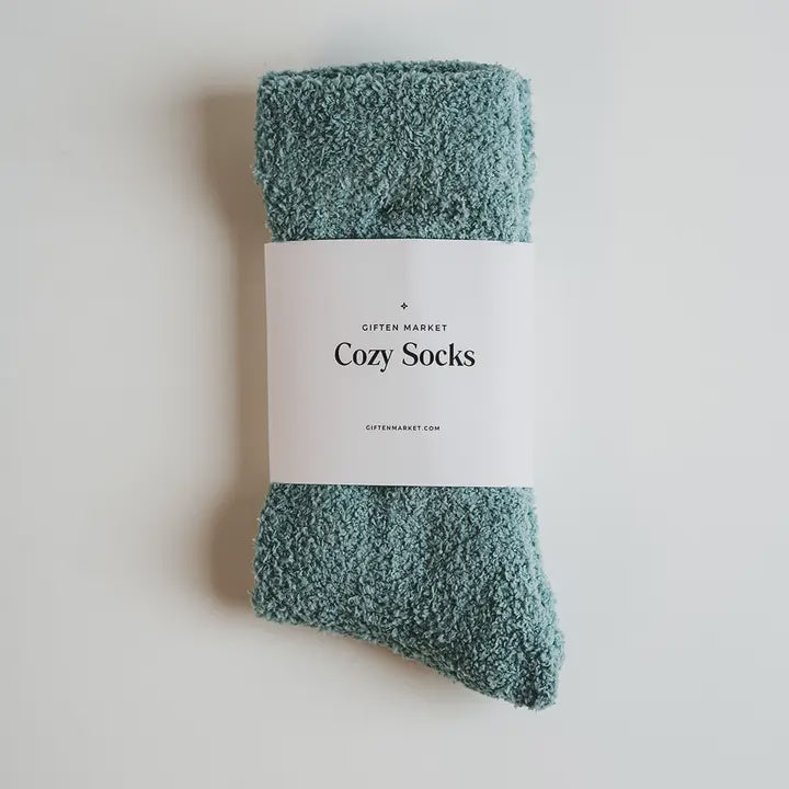 Product photograph by Giften Market of sage green fuzzy chenille socks featuring a white paper sleeve labeled Cozy Socks on a minimalist off-white background
