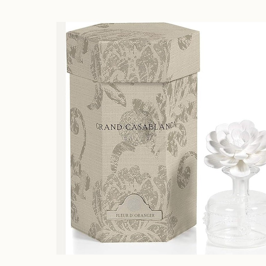Commercial product photography of a Grand Casablanca Fleur d’Oranger room diffuser set featuring a beige hexagonal damask-patterned box and a clear glass bottle with a white multi-layered ceramic flower topper against a minimalist studio background
