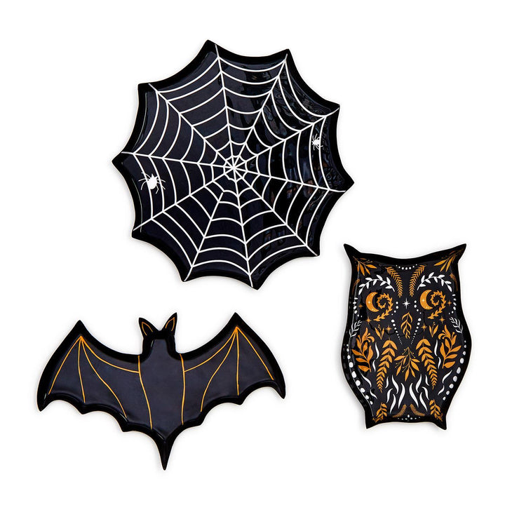 Ceramic Halloween-themed trinket dishes of a spiderweb, bat, and owl on a white background, featuring a glossy black glaze, delicate white spider web patterns, and golden folk-art celestial motifs on the owl