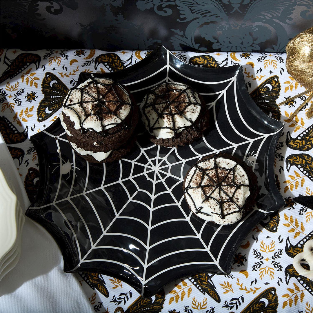 Photograph of three chocolate sandwich cookies with white spiderweb icing arranged on a glossy black spiderweb-shaped plate atop a decorative Halloween tablecloth featuring crow and moth motifs