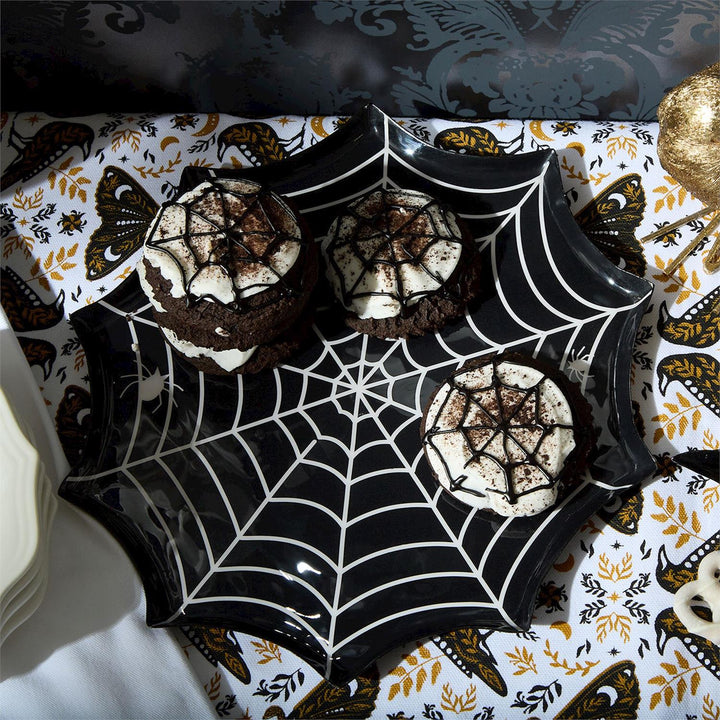 Photograph of three chocolate sandwich cookies with white spiderweb icing arranged on a glossy black spiderweb-shaped plate atop a decorative Halloween tablecloth featuring crow and moth motifs