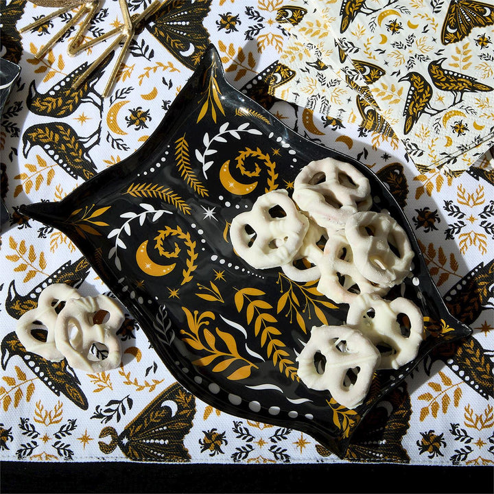 Lifestyle photograph of a Michel Design Works tray holding white chocolate covered pretzels on a tabletop, highlighting gold crescent moon motifs, white celestial vines, and a matching raven and moth patterned background