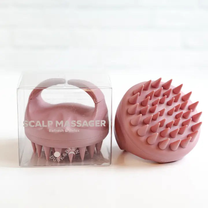 Product photograph of a Dusk & Dawn mauve silicone scalp massager featuring pointed flexible bristles and an ergonomic handle next to its clear retail packaging on a white surface