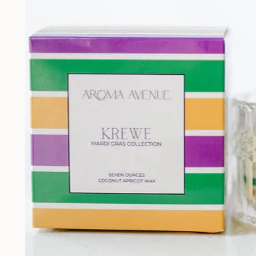 Product photograph by Aroma Avenue featuring a Krewe Mardi Gras Collection scented candle in an ornate cut-glass jar with a wooden wick alongside a packaging box with bold purple, green, and gold stripes on a minimalist white surface