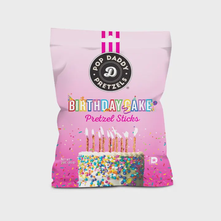 Commercial product photography for Pop Daddy Pretzels displays a light pink bag of Birthday Cake Pretzel Sticks featuring the black circular brand logo and a white frosted cake covered in multi-colored confetti sprinkles with several lit candles