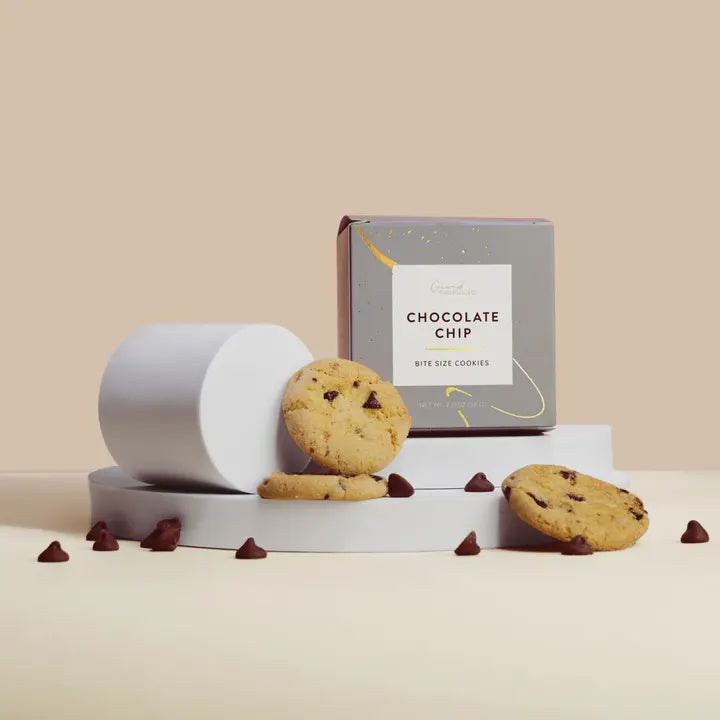 Product photograph of Grand bite-size chocolate chip cookies and their minimalist gray box with gold accents displayed on white geometric pedestals with scattered dark chocolate chips against a warm cream background