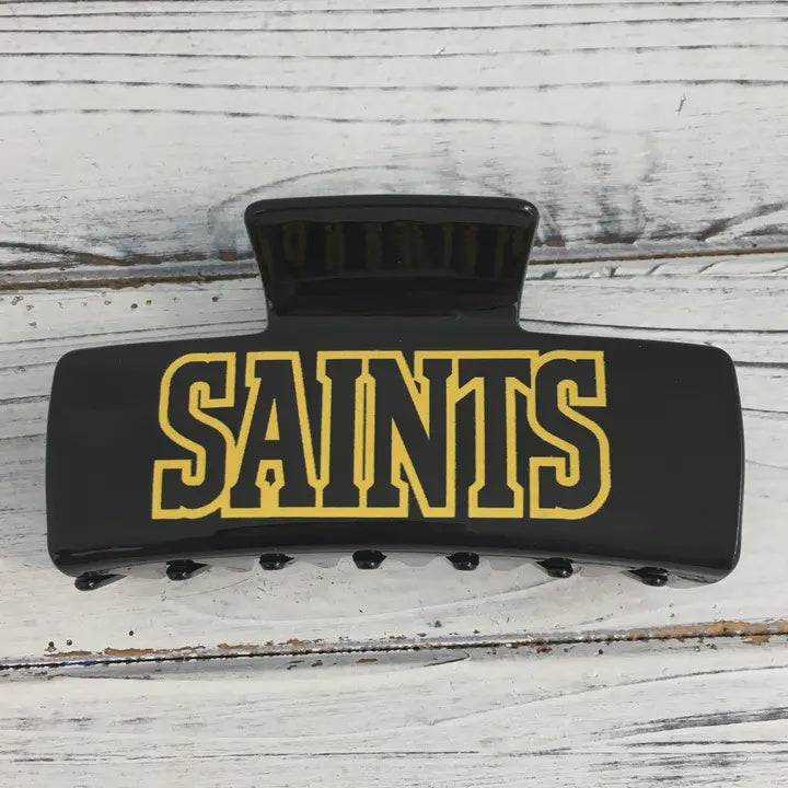 Acrylic hair claw clip featuring bold yellow varsity-style SAINTS lettering on a glossy black finish resting on a weathered white wooden surface