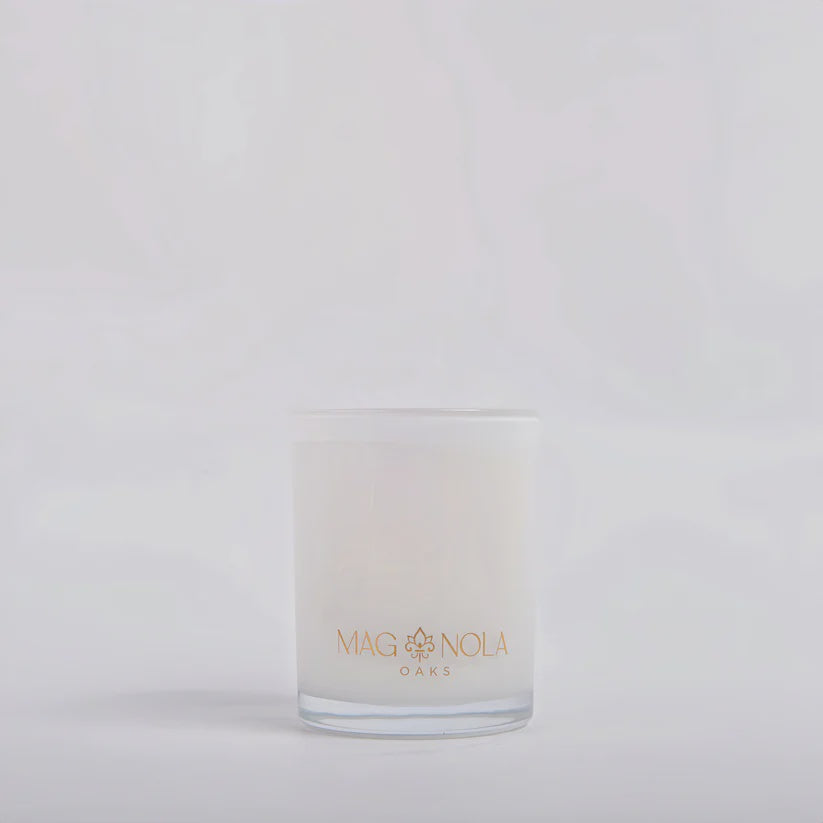 Product photograph by Magnolia Oaks of a minimalist white jar candle featuring an elegant gold fleur-de-lis logo and a thick clear glass base set against a clean white background