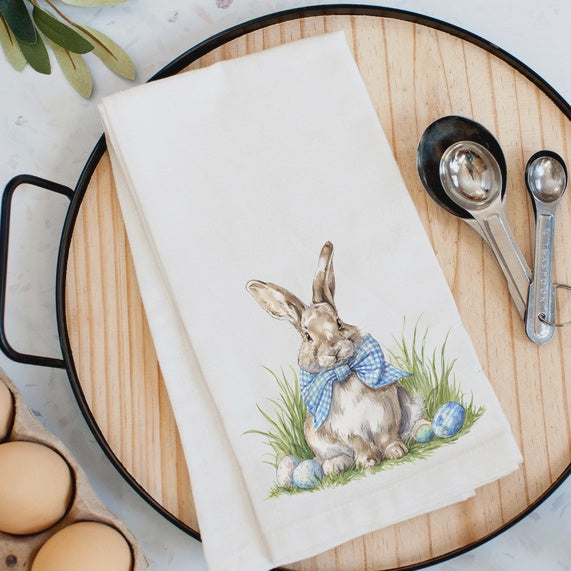 Watercolor-style illustration of a brown rabbit on a white tea towel laid over a circular wooden tray with measuring spoons and brown eggs, featuring a blue gingham bowtie and colorful Easter eggs tucked into blades of green grass