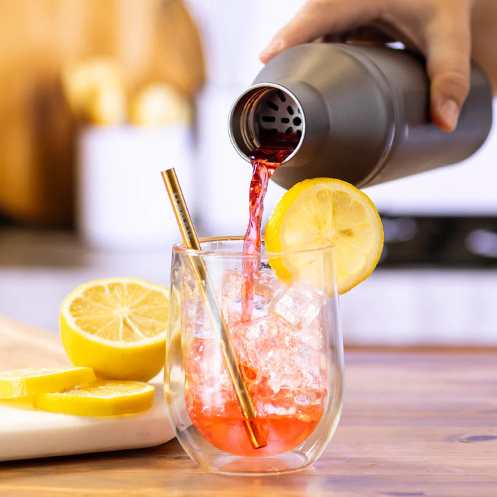 Photograph of a hand pouring a vibrant red drink from a matte charcoal cocktail shaker into a glass filled with ice cubes, a gold Contigo straw, and a bright yellow lemon wheel garnish on a wooden table