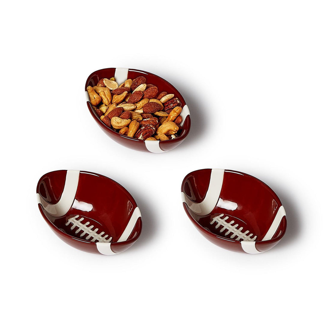 Glossy ceramic football-shaped snack bowls arranged on a white background featuring iconic white painted laces, a deep burgundy glaze, and one bowl filled with a variety of textured roasted nuts