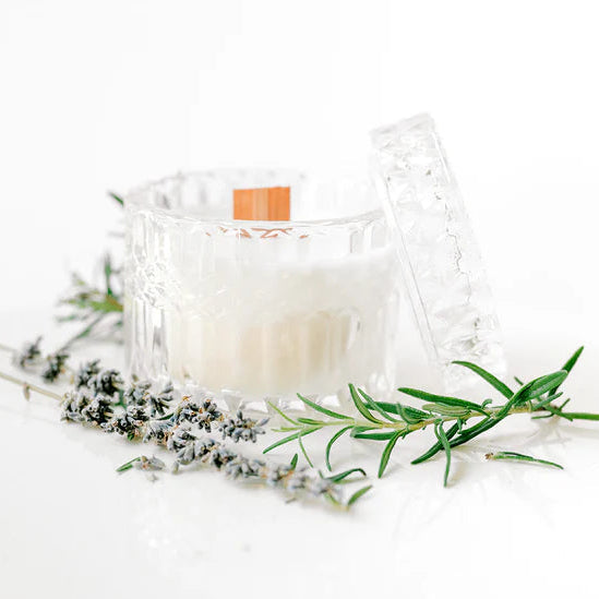 Still life photograph of a white candle in a faceted glass jar on a bright white surface featuring a wide wooden wick, vibrant green rosemary branches, and dried lavender sprigs