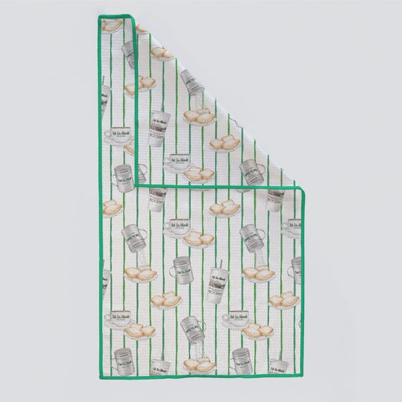 Illustrated waffle-weave kitchen towel featuring iconic Café Du Monde New Orleans motifs including plates of golden-brown beignets and silver powdered sugar shakers set against a green and white striped background