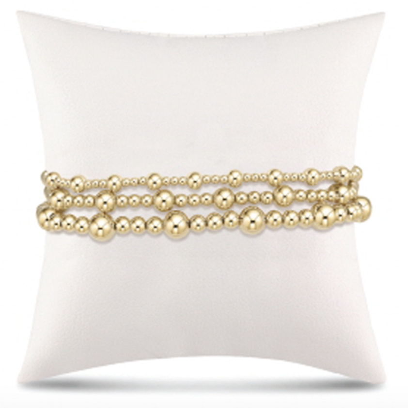 Product photograph of three tiered yellow gold beaded bracelets displayed on a white cushion featuring various sized polished beads and a high-shine reflective finish